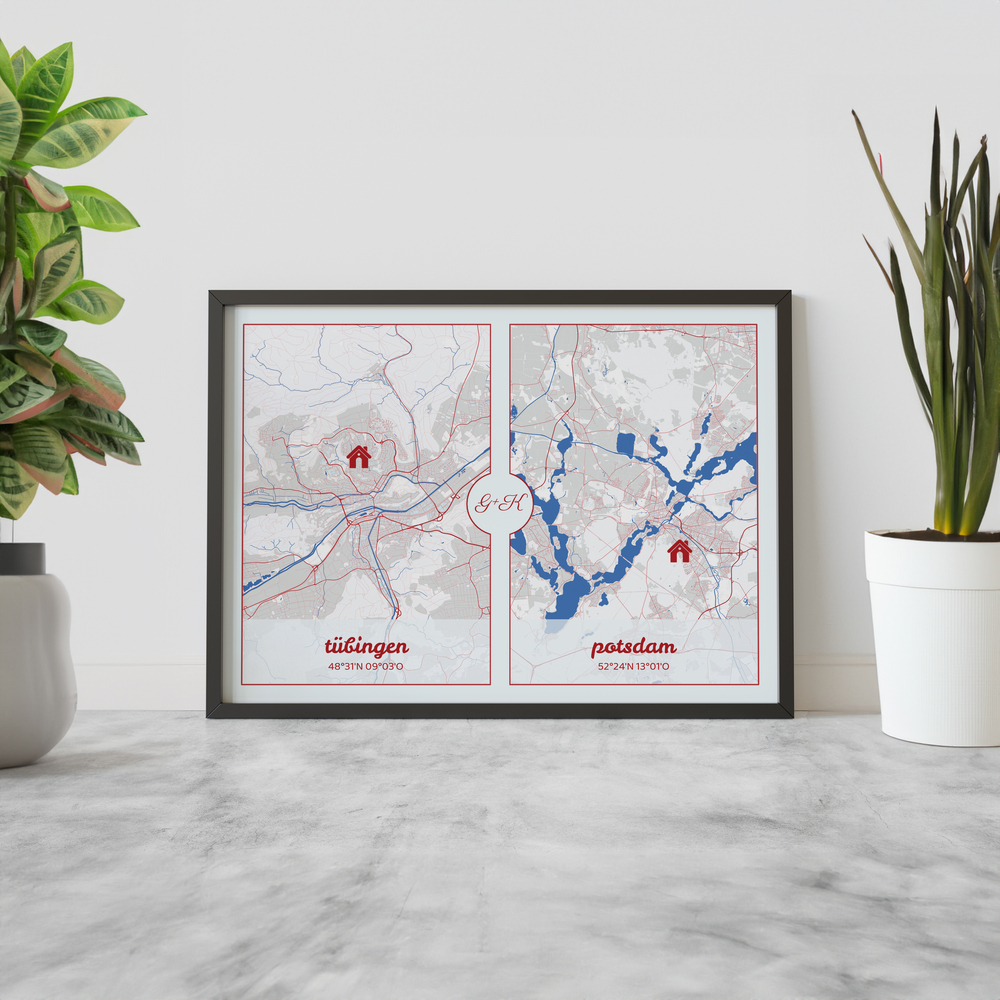 Mappe di due città con casette rosse in cornice nera, appoggiata a una parete chiara tra piante in vaso